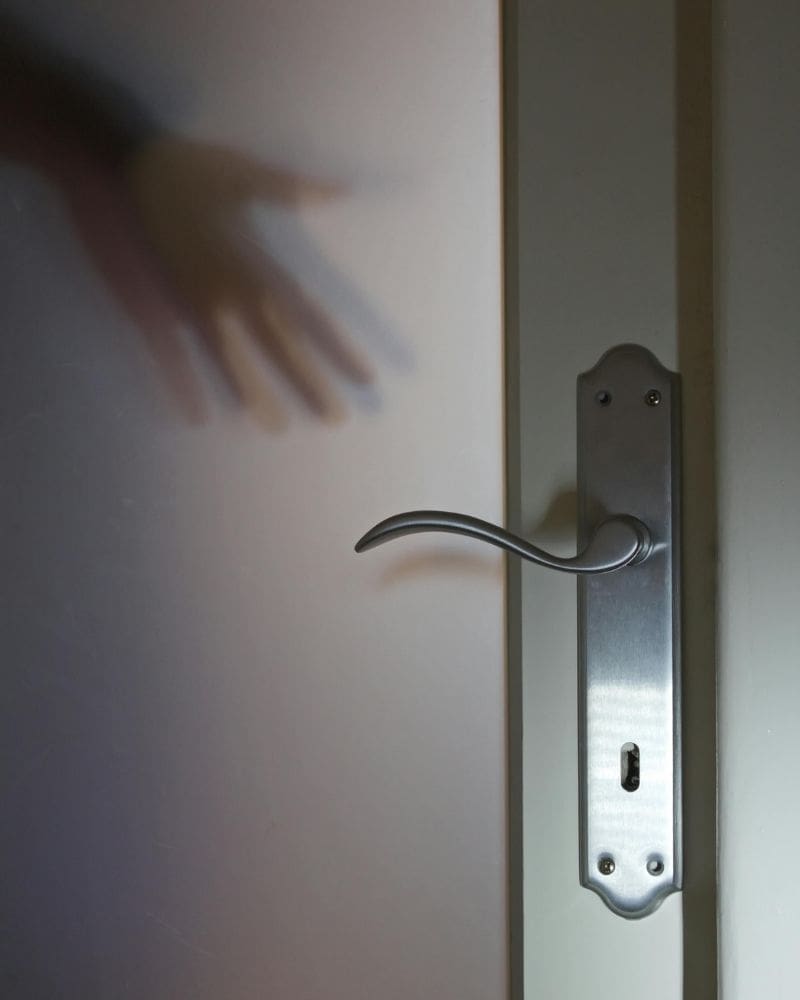 Outstretched hand behind a frosted door reaching toward a closed door handle with keyhole.